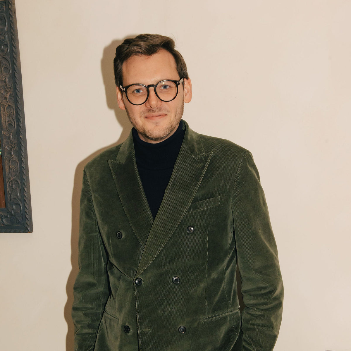 A man wearing a dark green velvet double-breasted blazer and matching trousers over a black turtleneck, standing in an indoor setting against a cream-colored wall.